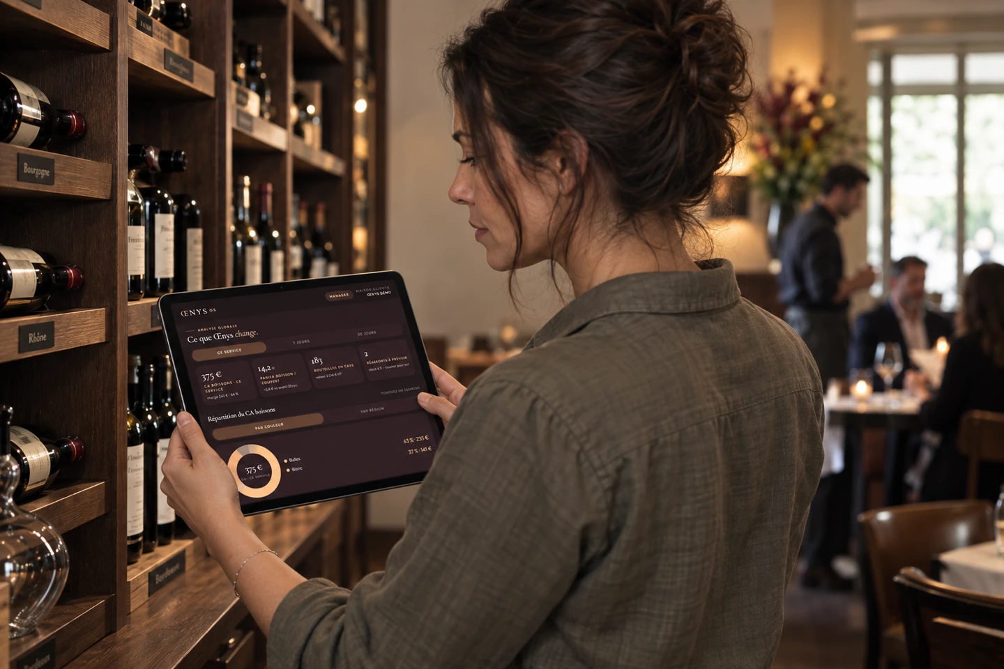 Scène illustrative : une restauratrice consulte Œnys OS sur sa tablette pour suivre sa cave, tandis que son équipe accompagne les clients en salle
